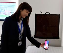 　無線給電システムの実証実験で、送電機（奥）の手前で受電機を取り付けたスマホを手にする担当者＝７月、愛知県稲沢市
