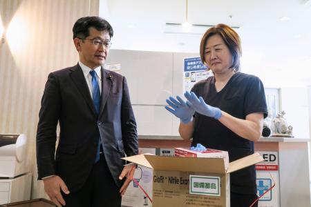 <p>　配送された国備蓄の医療用手袋を着ける「天王洲パークサイドビル歯科」の坂田香里院長。左は厚労省の仁木博文副大臣＝２３日午後、東京都品川区</p>