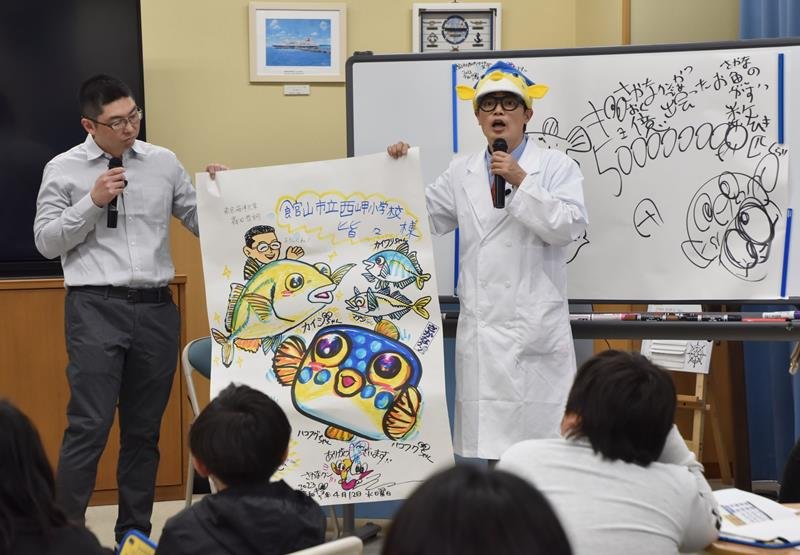 カイワリとマアジを掛け合わせたハイブリッド魚「カイジ」の絵を児童に披露するさかなクン（右）と森田准教授＝館山市
