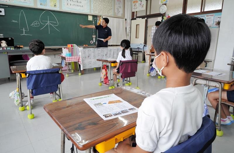 発案者の大谷代表の話を聞き、地元スギ材で作った机を使う１年生＝いすみ市立東小（写真を一部加工しています）
