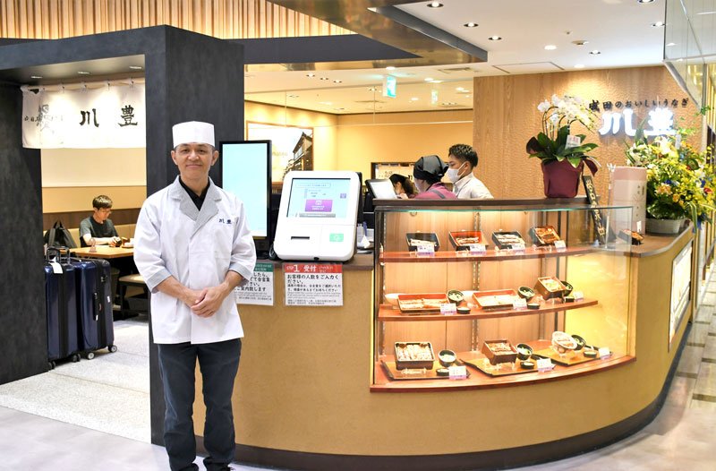 成田空港第１ターミナルにオープンした老舗うなぎ店「川豊」の店頭に立つ伊藤社長＝２２日、成田市