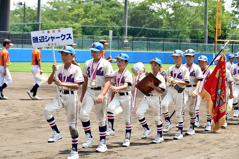 優勝旗などを手にグラウンドを行進する磯辺シャークスナイン＝２１日、横芝光町のふれあい坂田池公園球場