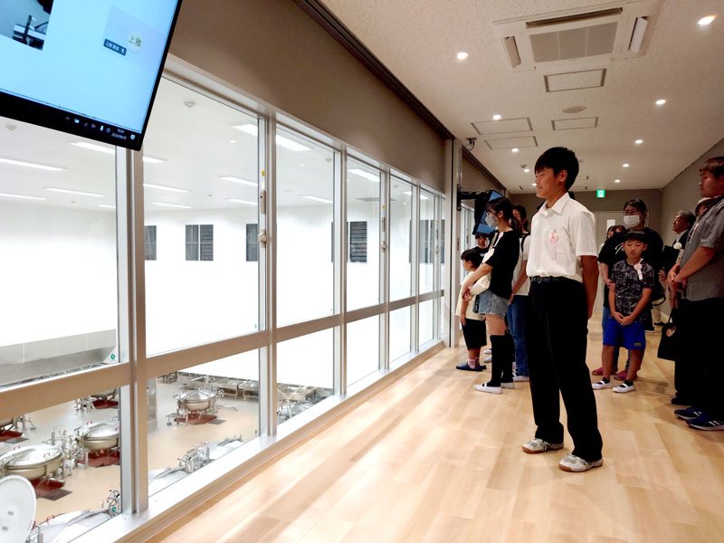 来月から稼働する市原学校給食センター見学エリアから説明を受けながら調理場を見学する市内小中学校の生徒児童ら＝２９日午前、市原市の市原学校給食センター