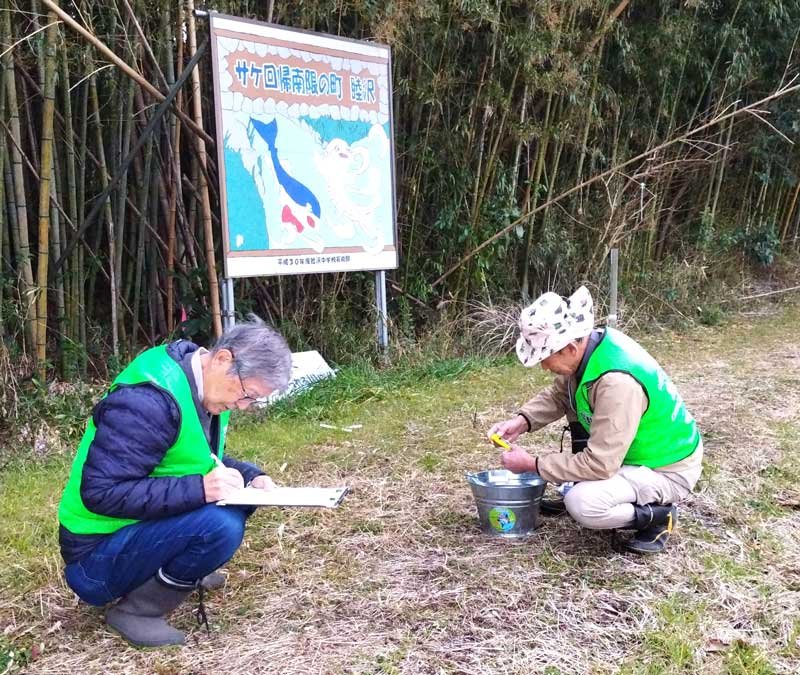 地元の中学生が作った看板の前で瑞沢川の水質調査をする松本さん（左）ら＝２月３日、睦沢町（松本さん提供）