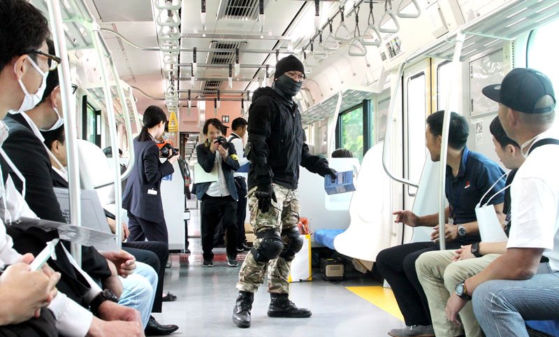 電車内にガソリンをまくしぐさを見せつける犯人役（中央）と、その様子を見る乗客役ら＝鴨川市
