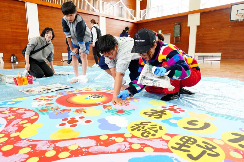 講師のバルサミコヤスさん（右）と一緒に手のひらをキャンバスの板に当てる児童＝勝浦市