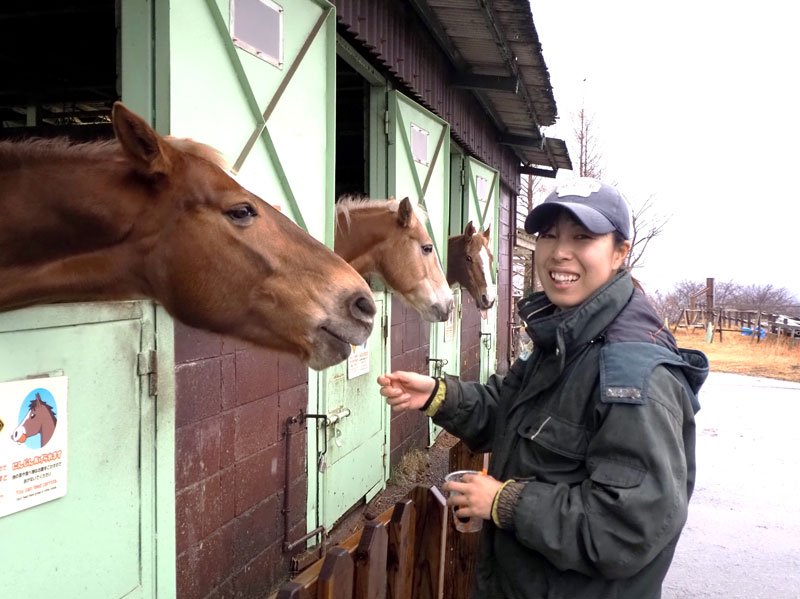 厩舎から顔を出す馬たちと国東さん。乗馬と合わせて餌やリ体験が楽しめる＝富津市
