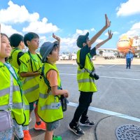 成田空港の駐機場から搭乗橋を歩く旅客に向けて手を振る小学生たち